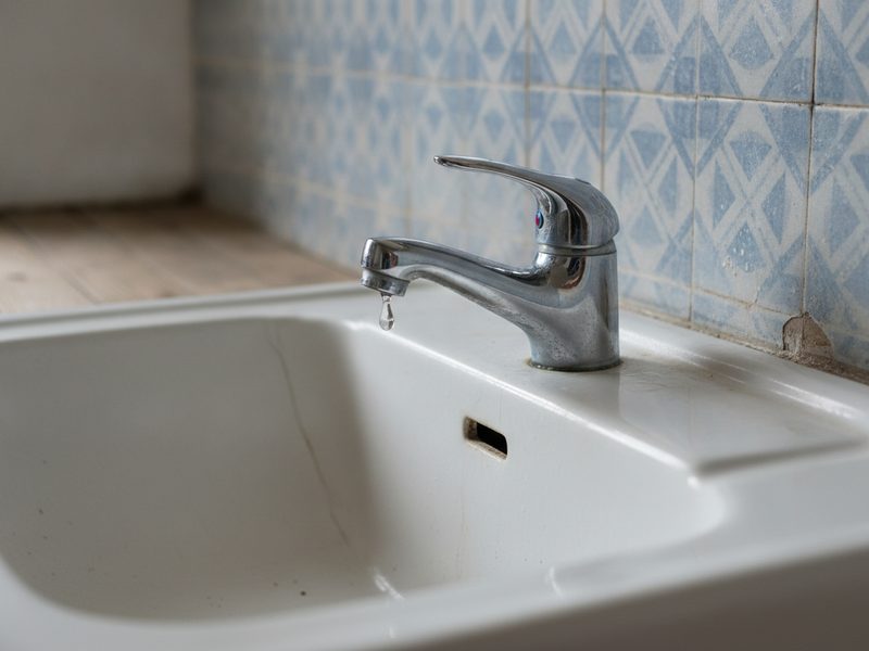 Mitigeur chromé monotrou sur un lavabo en céramique blanche, avec une goutte au bout du bec et un carrelage ancien à motifs derrière.